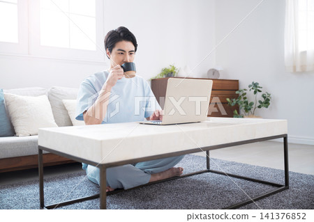Japanese man working or searching on his laptop; drinking coffee at home; full body shot of teleworking and relaxing. Japanese man working or searching on his laptop; drinking coffee at home; full body shot of teleworking and relaxing. 141376852