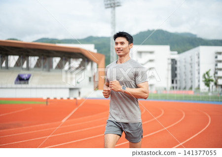 Asian male runner smiling while jogging on running track in sports stadium. Asian male runner smiling while jogging on running track in sports stadium. 141377055