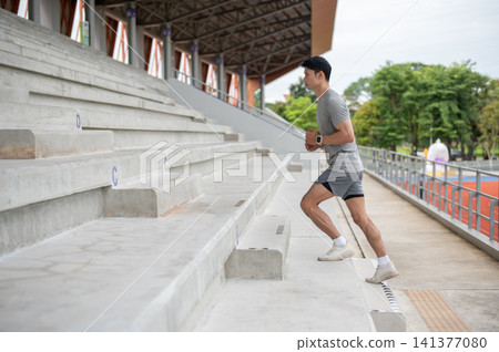 Fit Asian athletic man running up the concrete seating stairs before exercise in sports stadium. Fit Asian athletic man running up the concrete seating stairs before exercise in sports stadium. 141377080