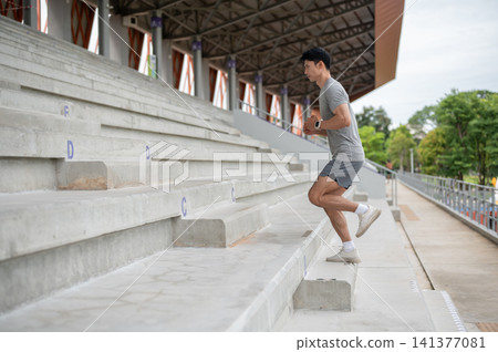 Active Asian athletic man sprinting upstairs during a workout session in sports stadium. Active Asian athletic man sprinting upstairs during a workout session in sports stadium. 141377081