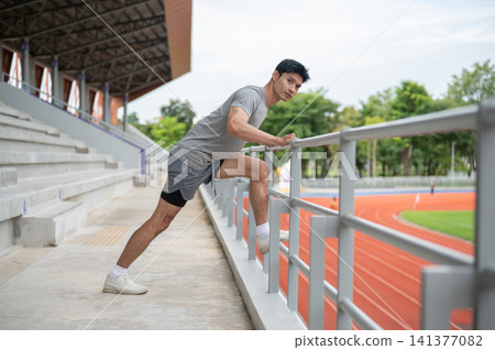 Athletic Asian man stretching leg muscles on a railing before exercise in sports stadium. Athletic Asian man stretching leg muscles on a railing before exercise in sports stadium. 141377082