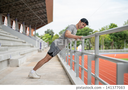 Asian athletic man stretching muscles against a railing before a running session in sports stadium. Asian athletic man stretching muscles against a railing before a running session in sports stadium. 141377083