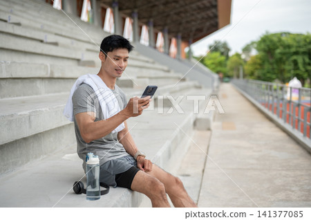 Asian athletic man sitting on seating stairs and using smartphone while resting in sports stadium. Asian athletic man sitting on seating stairs and using smartphone while resting in sports stadium. 141377085