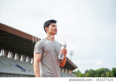 Thirsty Asian runner holding a water bottle after training in sports stadium. Thirsty Asian runner holding a water bottle after training in sports stadium. 141377125