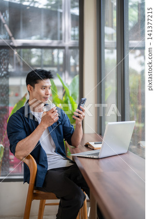 Asian freelancer man enjoy drinking coffee while using smartphone beside a laptop at table in cafe. Asian freelancer man enjoy drinking coffee while using smartphone beside a laptop at table in cafe. 141377215