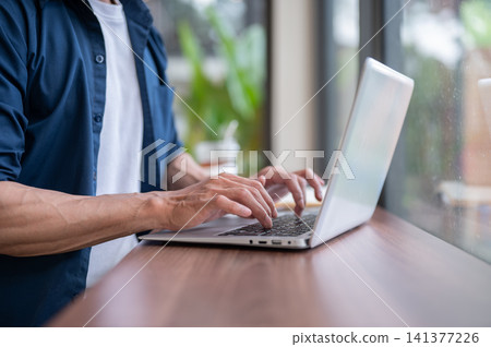Close up of asian man's hands typing on laptop at wooden table while working in the cafe. Close up of asian man's hands typing on laptop at wooden table while working in the cafe. 141377226