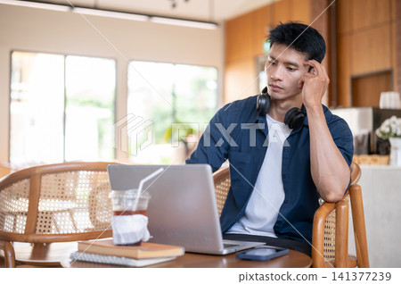 Asian man with headphones scratching temple and thinking while working on laptop at table in cafe. Asian man with headphones scratching temple and thinking while working on laptop at table in cafe. 141377239