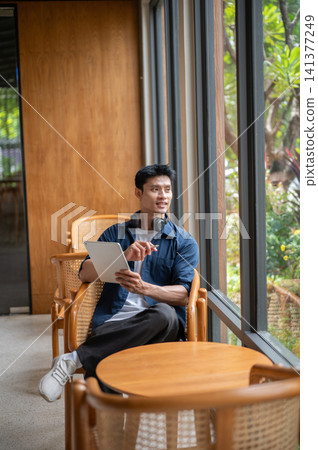 Asian freelancer man looking away from tablet while relaxing at wooden table in modern cafe. Asian freelancer man looking away from tablet while relaxing at wooden table in modern cafe. 141377249