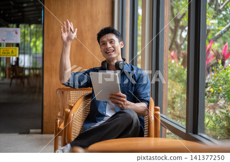 Asian man artist smiling while holding tablet and waving a hand while sitting at table in the cafe. Asian man artist smiling while holding tablet and waving a hand while sitting at table in the cafe. 141377250