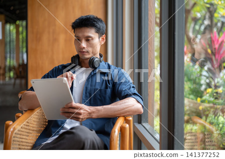 Asian man artist using tablet with stylus pen while working on a design at table the in cafe. Asian man artist using tablet with stylus pen while working on a design at table the in cafe. 141377252