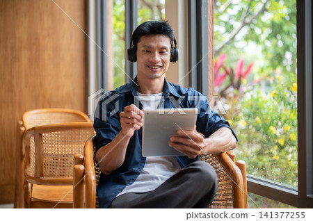 Asian artist man using a tablet and enjoy listening to music in headphones while working in the cafe Asian artist man using a tablet and enjoy listening to music in headphones while working in the cafe 141377255
