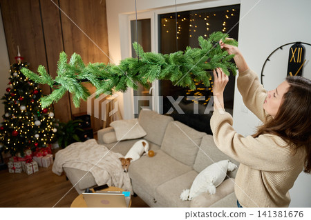 Woman Hanging Christmas Garland in Modern Living Room Woman Hanging Christmas Garland in Modern Living Room 141381676