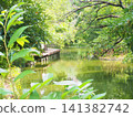 A lush green park with a pond and a wooden deck extending over the water's edge. 141382742