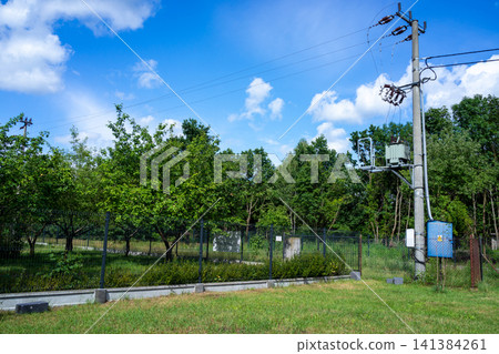 Electric utility pole with transformers, overhead power lines, fenced green landscape, trees, grass and clouds beneath bright blue sky, showing rural electrical infrastructure in daylight 141384261