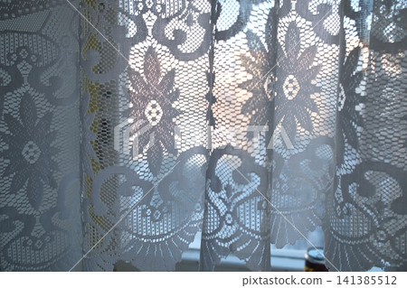 White lace curtains in window. Bright light through intricate floral design creates soft glow White lace curtains in window. Bright light through intricate floral design creates soft glow 141385512