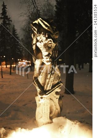 Tall stone sculpture of a draped figure brightly illuminated from below in a snow-covered park Tall stone sculpture of a draped figure brightly illuminated from below in a snow-covered park 141385514