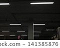 Linear lights on a dark industrial ceiling illuminate pipes, a vent, and a column in a modern space 141385876