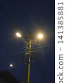 A utility pole with bright streetlights illuminates the dark night sky. 141385881