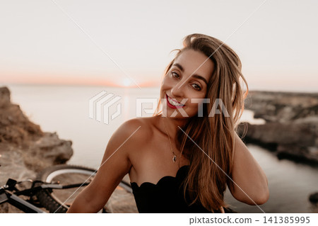 Woman sunset coast smiling young lady sitting on cliffs by the ocean during golden hour with a bicycle Woman sunset coast smiling young lady sitting on cliffs by the ocean during golden hour with a bicycle 141385995