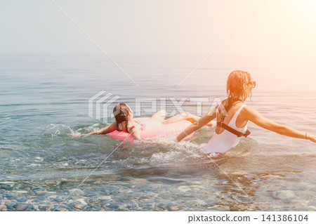 Mother daughter beach woman and girl playing in sea with pink inflatable ring during sunny summer vacation Mother daughter beach woman and girl playing in sea with pink inflatable ring during sunny summer vacation 141386104
