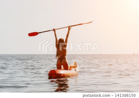 Woman Paddleboarding Ocean - A woman paddles on a stand-up paddleboard in the ocean. She is holding her paddle high in the air and smiling. 141386150