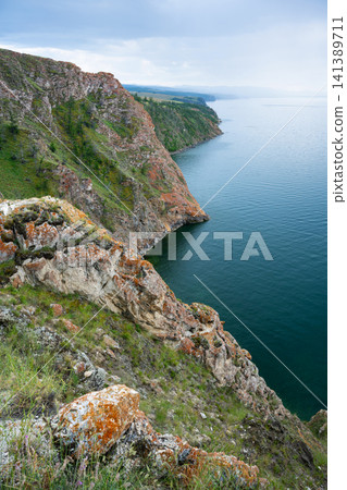 Vertical landscape of Lake Baikal, a steep rocky cliff covered in orange lichen dropping into deep blue water. Rugged coastline of Olkhon Island in summer 141389711