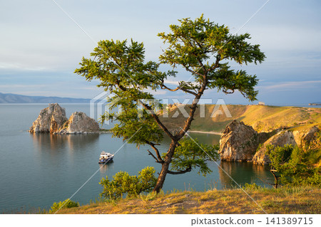 Landscape view of Lake Baikal with a prominent tree in the foreground. Shamanka Rock on Olkhon Island during golden hour. Summer travel destination in Siberia, Russia Landscape view of Lake Baikal with a prominent tree in the foreground. Shamanka Rock on Olkhon Island during golden hour. Summer travel destination in Siberia, Russia 141389715