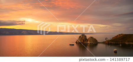 Panoramic sunset over Lake Baikal with Shamanka Rock. Vibrant orange sky and clouds reflecting on water. Scenic landscape of Olkhon Island in Russia Panoramic sunset over Lake Baikal with Shamanka Rock. Vibrant orange sky and clouds reflecting on water. Scenic landscape of Olkhon Island in Russia 141389717
