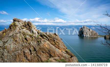 Rugged rock formation in the foreground overlooking Lake Baikal, Russia. Shamanka rock and mountains under a blue sky. Summer landscape of Olkhon island in Siberia Rugged rock formation in the foreground overlooking Lake Baikal, Russia. Shamanka rock and mountains under a blue sky. Summer landscape of Olkhon island in Siberia 141389719