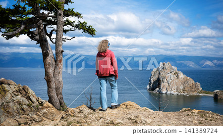 Female tourist looking at Shamanka Rock from a cliff at Olkhon Island on Lake Baikal, Russia. Woman in a red hoodie standing next to a pine tree. Summer travel destination Female tourist looking at Shamanka Rock from a cliff at Olkhon Island on Lake Baikal, Russia. Woman in a red hoodie standing next to a pine tree. Summer travel destination 141389724