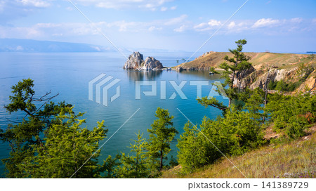 Wide landscape view of a blue Lake Baikal and prominent two-peaked rock formation. Green coniferous trees on a grassy cliff coast. Summer travel destination in nature Wide landscape view of a blue Lake Baikal and prominent two-peaked rock formation. Green coniferous trees on a grassy cliff coast. Summer travel destination in nature 141389729