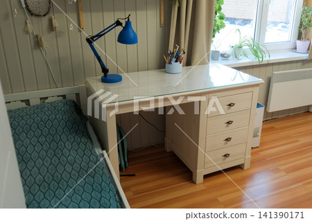 Bright school boy room with desk sunlit white desk beside bed, blue lamp, potted plants on windowsill, patterned bedspread, wooden floor, neat storage, calm study atmosphere. Learning environment Bright school boy room with desk sunlit white desk beside bed, blue lamp, potted plants on windowsill, patterned bedspread, wooden floor, neat storage, calm study atmosphere. Learning environment 141390171