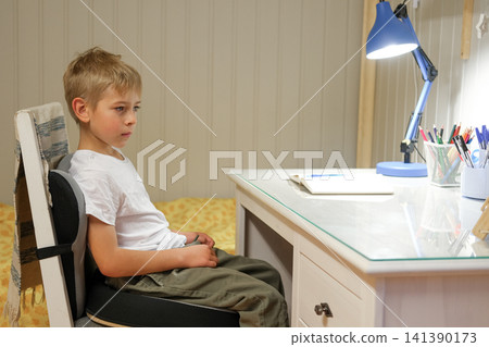 Boy kid sitting at study desk under lamp, hesitant expression while facing blank notebook and pencil cup, tidy bedroom, evening light creates quiet atmosphere, preparing homework. Child learning 141390173