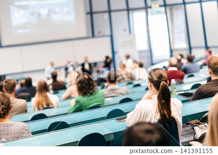 A large group of diverse people attending a professional presentation or educational workshop in a modern lecture hall. Focus on the audience from behind. A large group of diverse people attending a professional presentation or educational workshop in a modern lecture hall. Focus on the audience from behind. 141391155