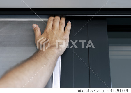 Hand pressing a white mosquito screen into the window frame, illustrating precise handwork and practical DIY maintenance. Hand pressing a white mosquito screen into the window frame, illustrating precise handwork and practical DIY maintenance. 141391789