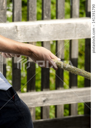 Person pulling a yellow braided rope against a wooden fence and foliage, illustrating the boat docking and mooring process. Person pulling a yellow braided rope against a wooden fence and foliage, illustrating the boat docking and mooring process. 141391790