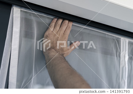 Hand securing a white mosquito screen into the window frame, illustrating precise handwork in home tasks and maintenance. Hand securing a white mosquito screen into the window frame, illustrating precise handwork in home tasks and maintenance. 141391793