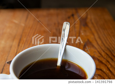 A closeup of a white ceramic cup filled with black coffee, with a metal spoon inside, placed on a rustic wooden table.Soft focus A closeup of a white ceramic cup filled with black coffee, with a metal spoon inside, placed on a rustic wooden table.Soft focus 141391798
