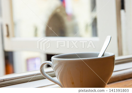 Closeup of a white ceramic coffee cup with a metal spoon, placed on a windowsill by an open window with a blurred old town of Tallinn in background. Soft focus Closeup of a white ceramic coffee cup with a metal spoon, placed on a windowsill by an open window with a blurred old town of Tallinn in background. Soft focus 141391803