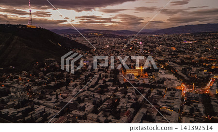 Aerial view of Tbilisi cityscape at dusk featuring Mtatsminda mountain with illuminated TV tower under a cloudy purple sunset sky in Georgia South Caucasus Aerial view of Tbilisi cityscape at dusk featuring Mtatsminda mountain with illuminated TV tower under a cloudy purple sunset sky in Georgia South Caucasus 141392514