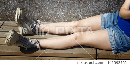 Young woman relaxes on a bench wearing shorts and sneakers on a sunny day Young woman relaxes on a bench wearing shorts and sneakers on a sunny day 141392731