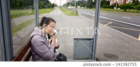 Woman waits at a bus stop while holding a small dog close in the city Woman waits at a bus stop while holding a small dog close in the city 141392755
