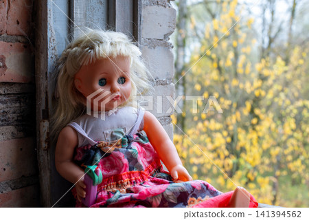 Blonde doll leans against brick window frame. Soft-focus yellow trees frame the background. Pink floral dress with green accents. Doll wears simple white top underneath Blonde doll leans against brick window frame. Soft-focus yellow trees frame the background. Pink floral dress with green accents. Doll wears simple white top underneath 141394562