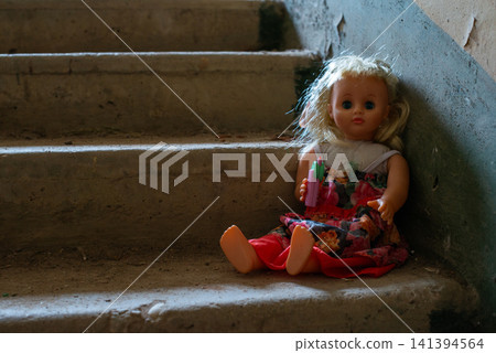 Doll sits alone on dusty concrete stairs. Blonde hair and colorful dress are visible. Soft lighting highlights worn, aged appearance. Staircase shows signs of neglect and time. Small figure adds eerie Doll sits alone on dusty concrete stairs. Blonde hair and colorful dress are visible. Soft lighting highlights worn, aged appearance. Staircase shows signs of neglect and time. Small figure adds eerie 141394564