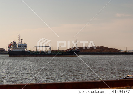 Large cargo ship floats calmly on wide water. Distant landmass forms soft silhouette behind vessel. Sky shows gentle gradient of warm evening light Large cargo ship floats calmly on wide water. Distant landmass forms soft silhouette behind vessel. Sky shows gentle gradient of warm evening light 141394577