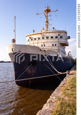 Old ship rests at water's edge, weathered and silent. Bare hull shows signs of time and tide's wear. Anchor chain drapes low, tethered to shore. Rusted rigging and mast stand tall against blue sky Old ship rests at water's edge, weathered and silent. Bare hull shows signs of time and tide's wear. Anchor chain drapes low, tethered to shore. Rusted rigging and mast stand tall against blue sky 141394584