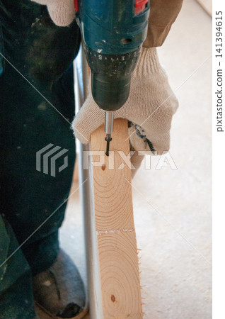 Hand holds drill, driving screw into wooden board. Work gloves protect hands during construction task. Wood grain visible, showing natural texture and knots Hand holds drill, driving screw into wooden board. Work gloves protect hands during construction task. Wood grain visible, showing natural texture and knots 141394615