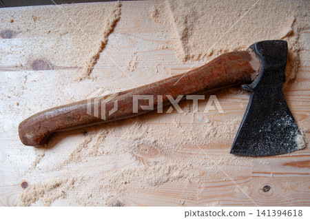 Rusty axe rests on wooden surface. Wood shavings surround the tool. Handle shows signs of wear. Blade has dark, weathered surface. Tool appears well-used and sturdy Rusty axe rests on wooden surface. Wood shavings surround the tool. Handle shows signs of wear. Blade has dark, weathered surface. Tool appears well-used and sturdy 141394618
