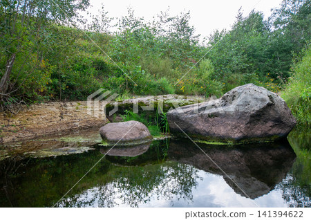 Still water reflects mossy rocks beside green forest. Vegetation grows thick on grassy bank. Large boulders rest at water's edge. Foliage and sky mirror in calm surface. Natural landscape shows quiet Still water reflects mossy rocks beside green forest. Vegetation grows thick on grassy bank. Large boulders rest at water's edge. Foliage and sky mirror in calm surface. Natural landscape shows quiet 141394622
