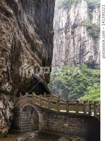 Where human heritage meets earth raw grandeur, a classic stone footbridge hugs the base of a monolithic rock wall within a deep limestone gorge. The weathered brickwork and elegant archway stand in Where human heritage meets earth raw grandeur, a classic stone footbridge hugs the base of a monolithic rock wall within a deep limestone gorge. The weathered brickwork and elegant archway stand in 141395221